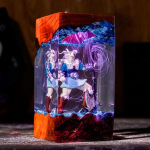May include: A rectangular block made of natural wood and clear resin, featuring a miniature warrior figurine inside. The warrior wears blue and white armor, holds a large axe, and is surrounded by swirling purple smoke effects within the resin.