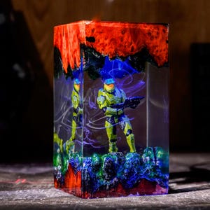 May include: A rectangular resin block featuring a green armored soldier figure holding a rifle inside. The top and bottom sections consist of vibrant orange and deep blue textured material, while the center contains swirling purple light effects.