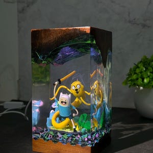 Peut inclure: Une lampe rectangulaire en résine avec une base et un sommet en bois, renfermant une scène miniature avec les personnages Finn et Jake. Le corps en résine transparente présente une figurine de chien jaune et un humain avec une épée.
