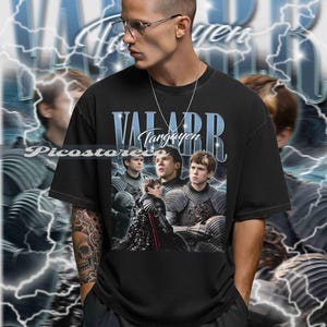 以下が含まれることがあります： ファンタジーシリーズのキャラクターが描かれた黒いTシャツ。青色の「VALARR Targaryen」の文字が特徴。鎧姿の人物と稲妻の背景がデザインされています。