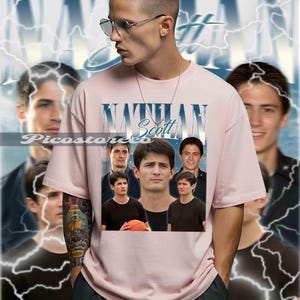 May include: A light pink t-shirt featuring the name "Nathan Scott" in a blue and white design. The shirt has multiple images of a person. The background has a lightning bolt design.