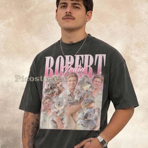 May include: A charcoal gray short-sleeve t-shirt featuring a graphic print of Robert Irwin holding several koalas. The text "ROBERT" appears in large pink letters at the top, with "Irwin" in a cursive script below. The shirt has a crew neckline.