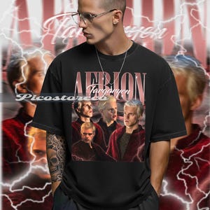 May include: Black t-shirt featuring a vintage-style graphic of a person with blonde hair and the text "Aemon Targaryen." The design includes a portrait of the character in a red jacket. The background has a lightning bolt design.