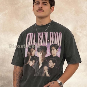 May include: A charcoal grey short-sleeve t-shirt featuring a graphic print of the name CHA EUN-WOO in pink lettering above a collage of four photographic portraits of the same individual against a purple and pink nebula background.