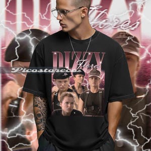 以下が含まれることがあります： ヴィンテージスタイルのグラフィックが特徴の黒いTシャツ。「Dizzy Flores」の文字と軍服姿の女性たちの画像がデザインされています。背景はピンクと白の稲妻模様です。