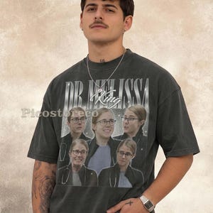 May include: A charcoal grey short-sleeve t-shirt featuring a vintage-style graphic print with the text "DR. MELISSA" and "King" in a stylised font. The design includes five repeated photographic portraits of a person wearing glasses and a stethoscope.