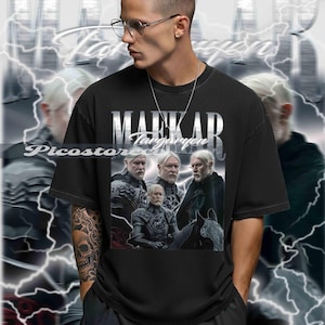 以下が含まれることがあります： 黒いTシャツ。前面には「MARKAR Targaryen」の文字が銀色のかすれたフォントでプリントされています。白い髪とひげの男性4人のグラフィックが描かれています。背景には稲妻のデザインがあります。