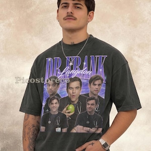 Op de afbeelding: Een antracietgrijs t-shirt met korte mouwen en een grafische print van Dr. Frank Langdon. Het ontwerp bevat de tekst "DR. FRANK Langdon" in paars en wit, samen met vijf fotografische afbeeldingen van het personage met een stethoscoop.