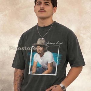 Könnte beinhalten: Ein anthrazitfarbenes Kurzarm-T-Shirt mit einem rechteckigen Grafikdruck von Johnny Depp, der einen Hut und ein weißes T-Shirt trägt. Der Druck enthält einen blauen Pinselstrich-Hintergrund und den Text Johnny Depp. Das Wort Picostore ist sichtbar.