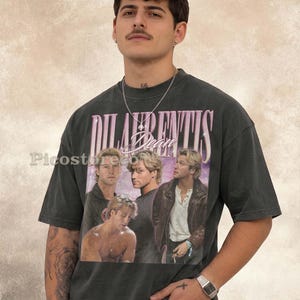 May include: A charcoal gray short-sleeve t-shirt featuring a graphic print of a person in various poses. The text "DILAURENTIS" appears in large pink letters at the top, with the name "Dean" in a cursive script below it. The shirt has a crew neckline.