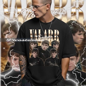 以下が含まれることがあります： 金色の「VALARR Targaryen」のグラフィックと画像のコラージュが施された黒いTシャツ。背景には、稲妻のアクセントが付いた金色のデザインがあります。画像内の人物はメガネとシルバーのチェーンを着用しています。
