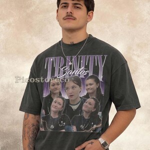 Könnte beinhalten: Ein anthrazitfarbenes Kurzarm-T-Shirt mit einem Grafikdruck im Vintage-Stil. Das Design zeigt das Wort TRINITY in großen violetten Buchstaben, den Namen Santos in weißer Schreibschrift und eine Collage aus fünf Fotos derselben Person.