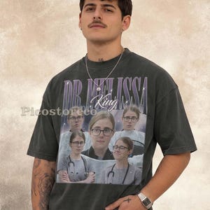 Może przedstawiać: Ciemnoszary t-shirt z krótkim rękawem z nadrukiem graficznym zawierającym napis DR. MELISSA oraz King. Wzór przedstawia kilka zdjęć osoby w okularach i ze stetoskopem na tle rozgwieżdżonego, kosmicznego nieba.