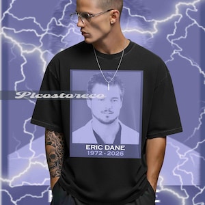 May include: Black t-shirt featuring a portrait of Eric Dane with the text "ERIC DANE 1972-2026" in a purple rectangle. The shirt is worn by a person with glasses and a silver necklace, set against a purple lightning background.