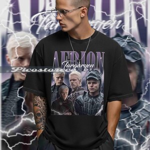 以下が含まれることがあります： 黒いTシャツ。紫と白で「Aemon Targaryen」の文字と画像のコラージュ。白い髪と厳しい表情の男性の画像が含まれています。背景には稲妻のデザインがあります。
