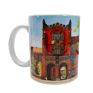 Puede incluir: Taza de cer&aacute;mica blanca con una ilustraci&oacute;n colorida de un edificio de ladrillo rojo con puertas arqueadas y ventanas. El edificio tiene un escudo azul y dorado sobre la entrada.