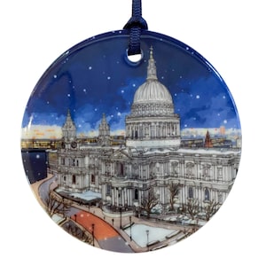 Puede incluir: Un adorno redondo de cerámica azul y blanco con una escena nevada de la Catedral de San Pablo en Londres. La catedral está representada en detalle, con su icónica cúpula y aguja. El adorno es perfecto para añadir un toque de encanto londinense a tu árbol de Navidad.