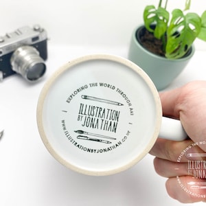 Puede incluir: Taza de cer&aacute;mica blanca con una ilustraci&oacute;n en blanco y negro de l&aacute;pices y pinceles. El texto en la taza dice "Explorar el mundo a trav&eacute;s del arte. Ilustraci&oacute;n de Jonathan. www.illustrationbyjonathan.co.uk"