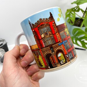 Puede incluir: Taza de cer&aacute;mica blanca con una ilustraci&oacute;n colorida de un edificio de ladrillo rojo con puertas arqueadas y ventanas. El edificio tiene un techo azul y est&aacute; rodeado de &aacute;rboles verdes. La taza tiene un asa negra.