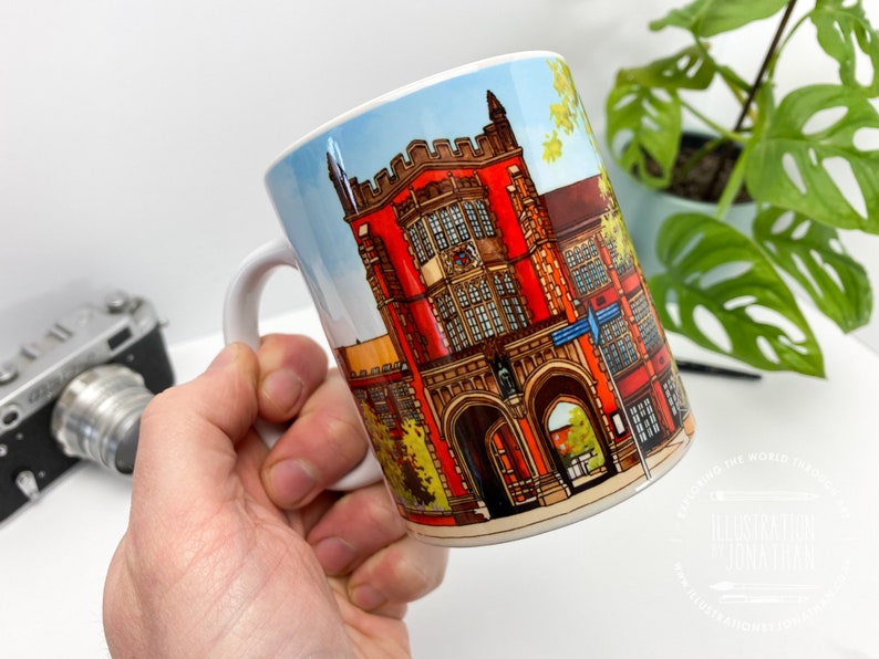 Puede incluir: Taza de cer&aacute;mica blanca con una ilustraci&oacute;n colorida de un edificio de ladrillo rojo con puertas arqueadas y ventanas. El edificio tiene un techo azul y est&aacute; rodeado de &aacute;rboles verdes. La taza tiene un asa negra.