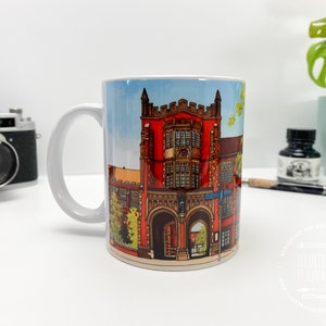 Puede incluir: Taza de cer&aacute;mica blanca con una ilustraci&oacute;n colorida de un edificio de ladrillo rojo con puertas arqueadas y ventanas. El edificio est&aacute; rodeado de &aacute;rboles verdes y un cielo azul.