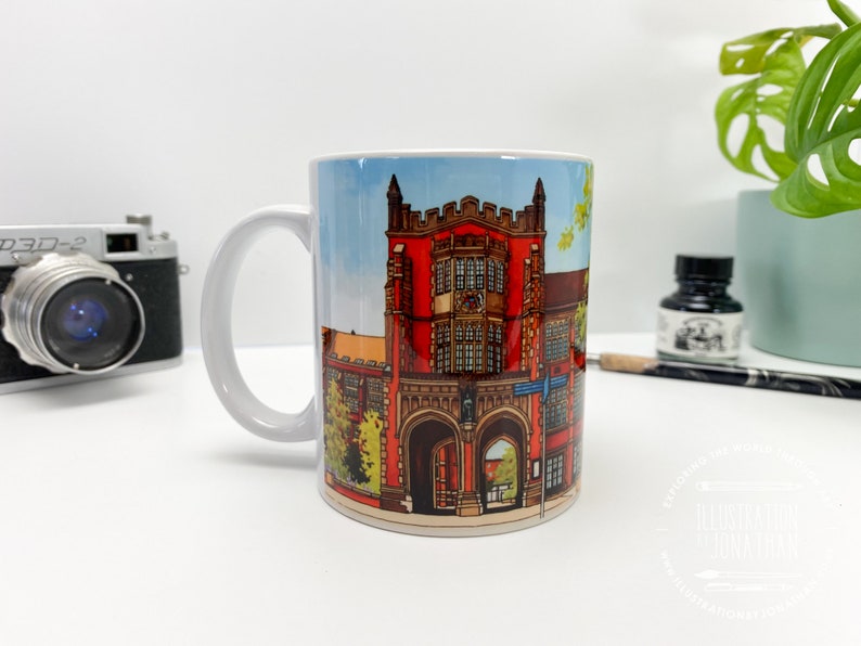 Puede incluir: Taza de cer&aacute;mica blanca con una ilustraci&oacute;n colorida de un edificio de ladrillo rojo con puertas arqueadas y ventanas. El edificio est&aacute; rodeado de &aacute;rboles verdes y un cielo azul.