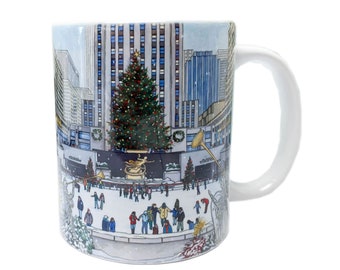 May include: White ceramic mug with a colorful illustration of a winter scene in New York City. The illustration features the Rockefeller Center Christmas tree, ice skaters, and the iconic building.