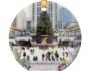 May include: A round ceramic ornament featuring a winter scene of a decorated Christmas tree in front of a skyscraper in a city. People are ice skating on a rink in the foreground. The ornament is white with a gold ribbon.