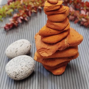 Puede incluir: Una torre de piedras planas de color naranja-marrón, con dos piedras lisas grises a la izquierda. El fondo presenta una superficie gris con líneas verticales y follaje rojo y verde borroso.
