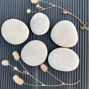 Puede incluir: Cinco piedras de río lisas de color gris claro dispuestas sobre una superficie gris oscuro con estrías. Se colocan hierbas secas de color beige alrededor de las piedras, añadiendo una estética natural. Las piedras varían en tamaño y forma.