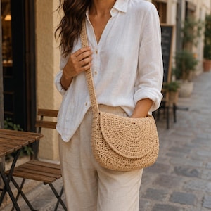 Puede incluir: Un bolso bandolera de ganchillo en color beige con solapa redondeada y correa tejida a juego. El bolso presenta un patrón de puntos circulares texturizados en todo el cuerpo y la solapa, lo que le confiere un aspecto estructurado y flexible.