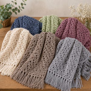 May include: A collection of six crocheted blankets featuring an openwork pattern and fringed edges. The blankets are displayed in various colors including cream, dark blue, sage green, mauve, brown, and gray, all arranged in a layered pile.