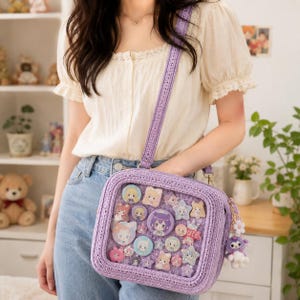 May include: A square, lavender-colored crochet crossbody bag featuring a clear front display window filled with various circular and star-shaped character pins. The bag includes a matching crochet shoulder strap and a small plush penguin charm.