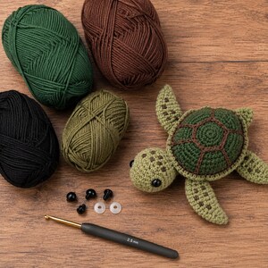 Op de afbeelding: Een gehaakte schildpad met een groen en bruin schild, vier bollen garen in donkergroen, bruin, zwart en olijf, vier kleine zwarte plastic veiligheidsoogjes met twee doorzichtige ringetjes en een zwarte haaknaald met 2,5 mm maat.