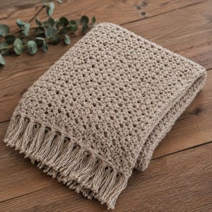 May include: A beige crochet throw blanket featuring a repeating openwork stitch pattern and a decorative fringe along the edge. The textile has a textured, knotted appearance and is folded into a rectangular shape with visible yarn loops.