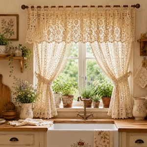 May include: A cream-colored crochet curtain set featuring an intricate floral pattern and a scalloped bottom edge, suspended from a wooden rod. The curtain is divided into two panels tied back to reveal a window with potted herbs on the sill.