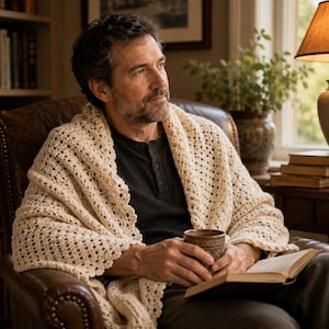 May include: A cream-colored, open-weave crochet shawl draped over the shoulders. The shawl features a delicate, repeating geometric pattern with a scalloped edge, providing a textured, lightweight layer over a dark-colored shirt.