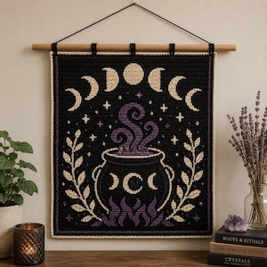 May include: A rectangular crochet wall hanging featuring a black background with a central black cauldron emitting purple swirling steam. The design includes cream-colored moon phases, stars, and leafy branches, all suspended from a wooden rod.