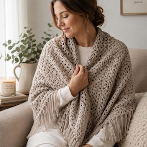 May include: A beige crochet shawl featuring an intricate openwork pattern and long fringe trim along the bottom edge. The shawl is draped over the shoulders, showcasing the textured yarn construction and the consistent stitch design throughout.