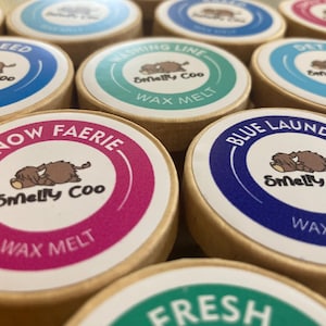 May include: A selection of wax melt containers with circular labels. The labels display the text "Smelly Coo" and a cartoon animal, with names such as "Snow Faerie" and "Blue Laundry." The containers are round with a natural wood finish. Measurements are not applicable.