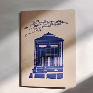 Puede incluir: Una ilustración azul de una casa con un porche y escalones sobre un fondo de papel marrón. La casa tiene una puerta y ventanas blancas. Hay flores azules sobre la casa.