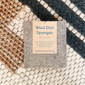 May include: Two gray wool dish sponges, 5 inches by 5 inches, with a brown paper label that reads "Wool Dish Sponges, QTY. 2, 5 x 5", "Made in New Orleans | 100% wool felt, 100% recycled paper packaging, Donates 10% of proceeds to The Wetlands Initiative", and "GOODS THAT MATTER".