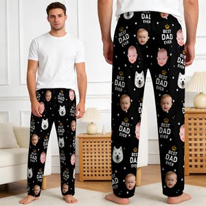 Puede incluir: Pantalones de pijama negros con un patrón repetido de caras de bebé, estrellas blancas y el texto "BEST DAD EVER" en blanco. Una cara de perro blanco y una corona dorada también forman parte del diseño. Los pantalones son usados por una persona que lleva una camiseta blanca.