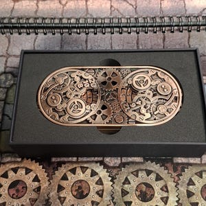 Può includere: Una fibbia per cintura in stile steampunk color rame con intricati disegni di ingranaggi, presentata in una scatola nera. La fibbia è di forma ovale e presenta una complessa disposizione di ingranaggi interconnessi. Lo sfondo include una superficie a tema fantasy.