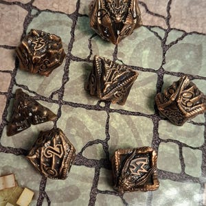 Puede incluir: Un conjunto de dados poliédricos de color bronce, incluyendo D4, D6, D8, D10, D12 y D20, dispuestos sobre un tablero de juego. Los dados tienen diseños intrincados y son adecuados para juegos de rol de mesa.