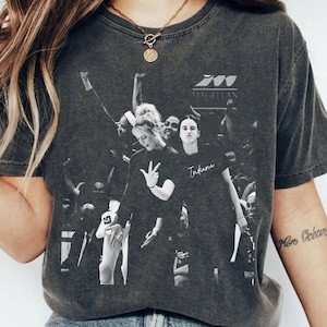 Pode incluir: T-shirt cinzento escuro com uma imagem a preto e branco de um grupo de pessoas. A palavra "Indiana" está impressa na t-shirt. A t-shirt tem gola redonda e mangas curtas. A t-shirt é feita de um material macio e confortável.