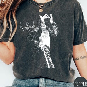 May include: A charcoal gray short-sleeve t-shirt featuring a white graphic print of Michael Jackson in a fedora and sequined jacket, with his signature printed above the figure. The shirt has a crew neckline and a faded, vintage-style wash.