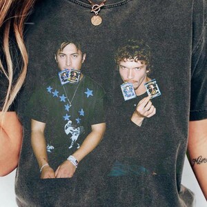 May include: Dark gray t-shirt featuring a graphic of two men holding cards. The shirt has a vintage, worn look. One man's shirt has blue star and figure graphics. The other man's shirt is plain. The shirt is paired with blue jeans.