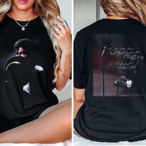 May include: Black t-shirt featuring a portrait of a musician with a hat. The back of the shirt displays a microphone, a hat on a stool, and handwritten text. The shirt is a comfortable, casual wear item.
