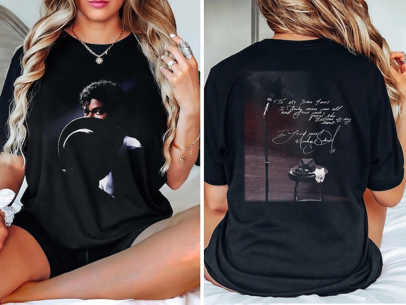 May include: Black t-shirt featuring a portrait of a musician with a hat. The back of the shirt displays a microphone, a hat on a stool, and handwritten text. The shirt is a comfortable, casual wear item.